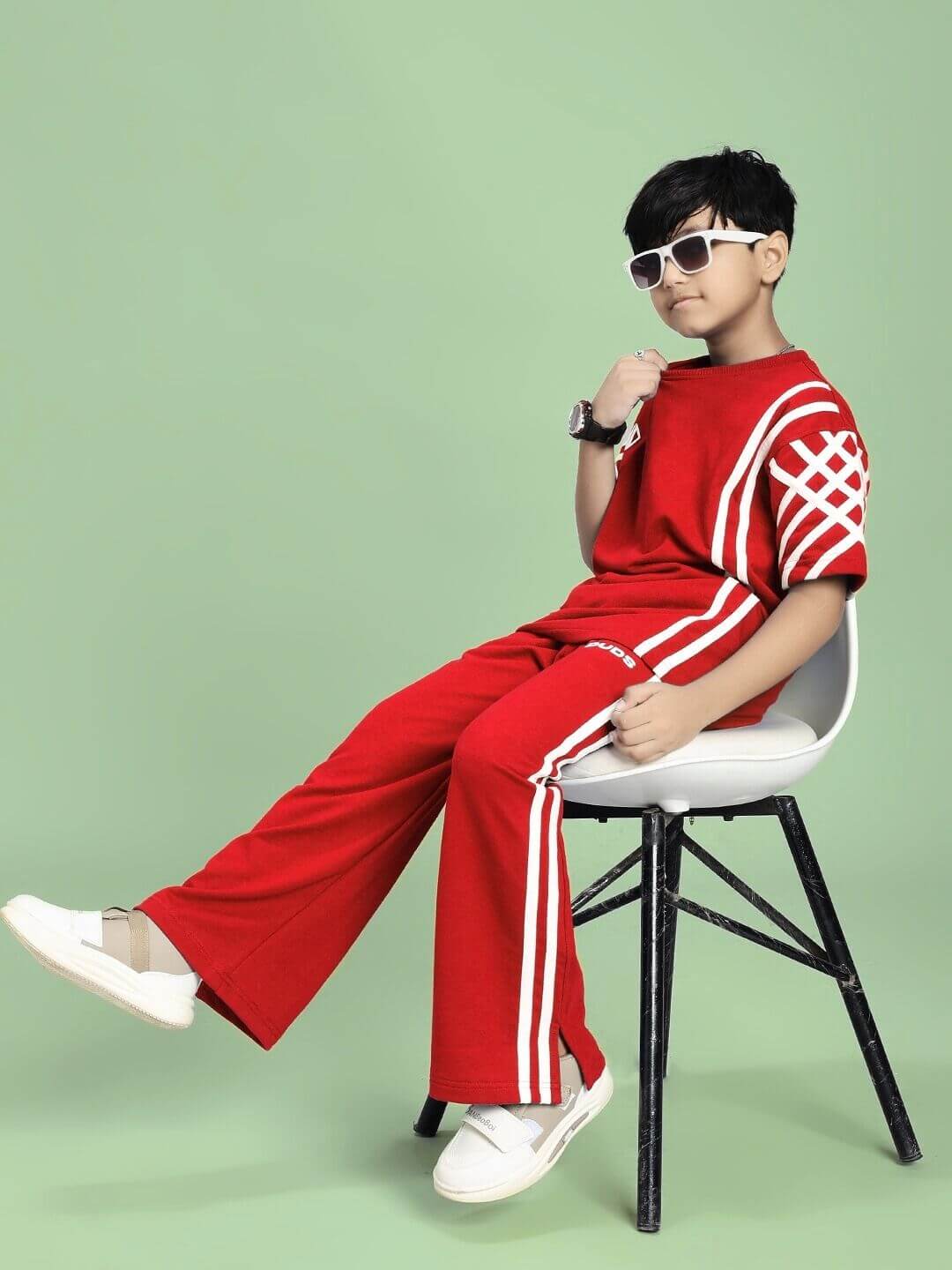 Child wearing UNITED STRIPE T-SHIRT FOR BOYS AND GIRLS RED with sporty stripe detail, sitting on a chair against a green background.