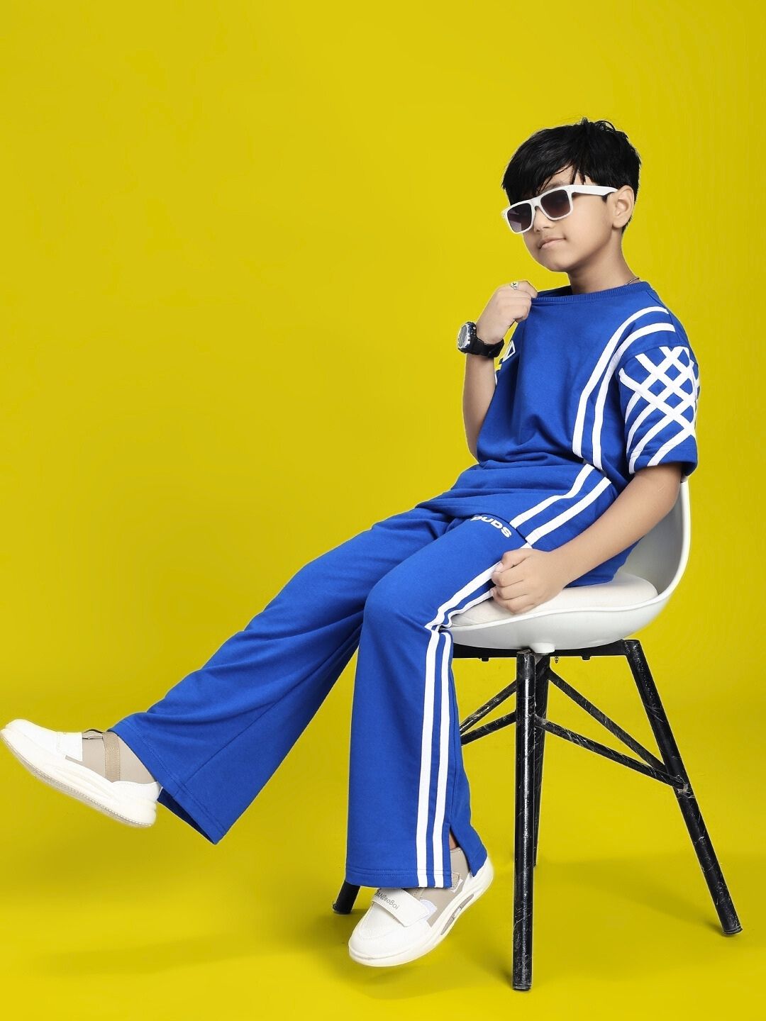Child wearing UNITED STRIPE T-SHIRT FOR BOYS AND GIRLS BLUE with matching pants and white shoes, sitting on a chair against a yellow background.