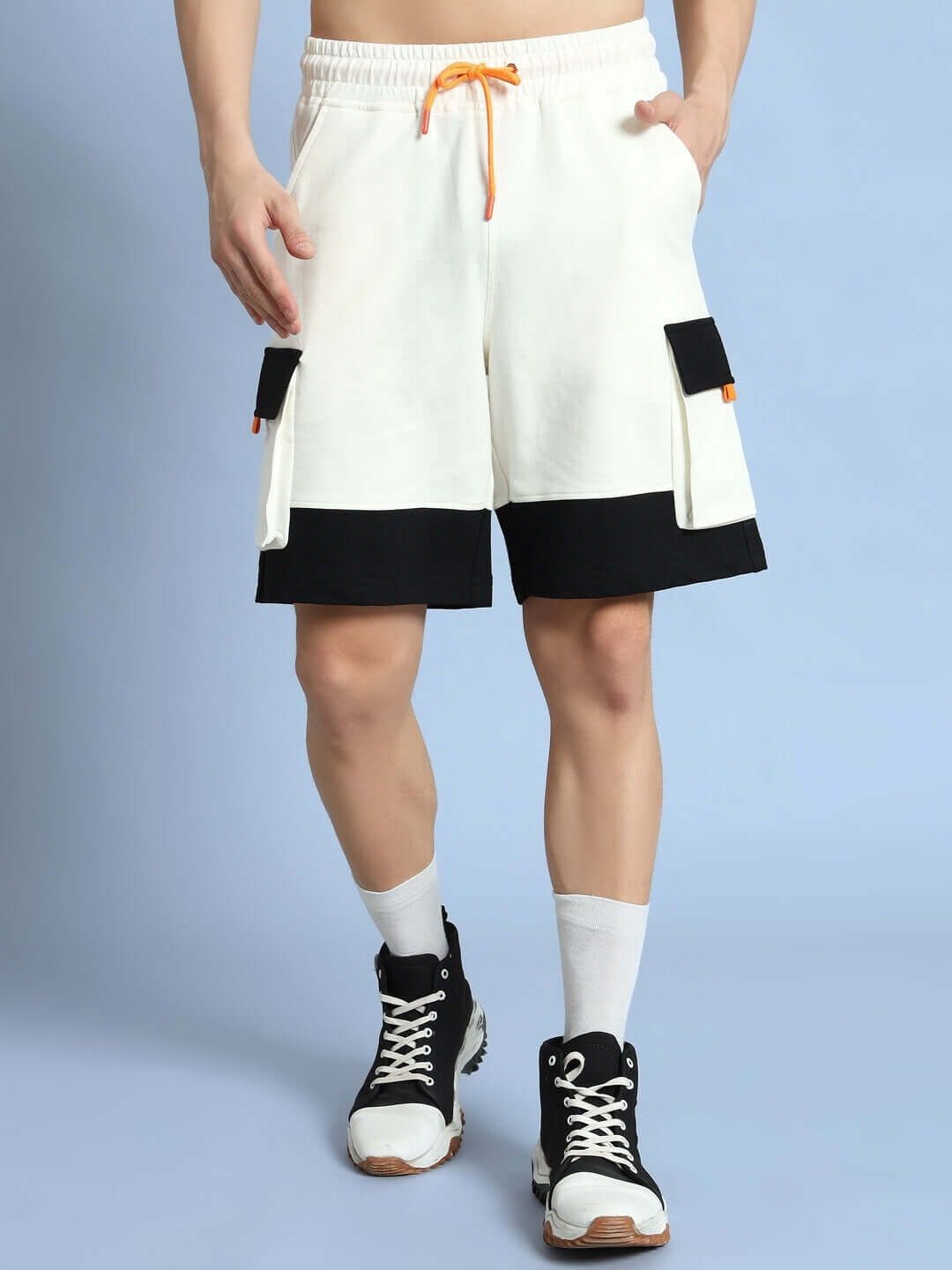 Man wearing off-white and black utility cargo shorts with pockets, featuring an orange drawstring, modeled with black sneakers and white socks.