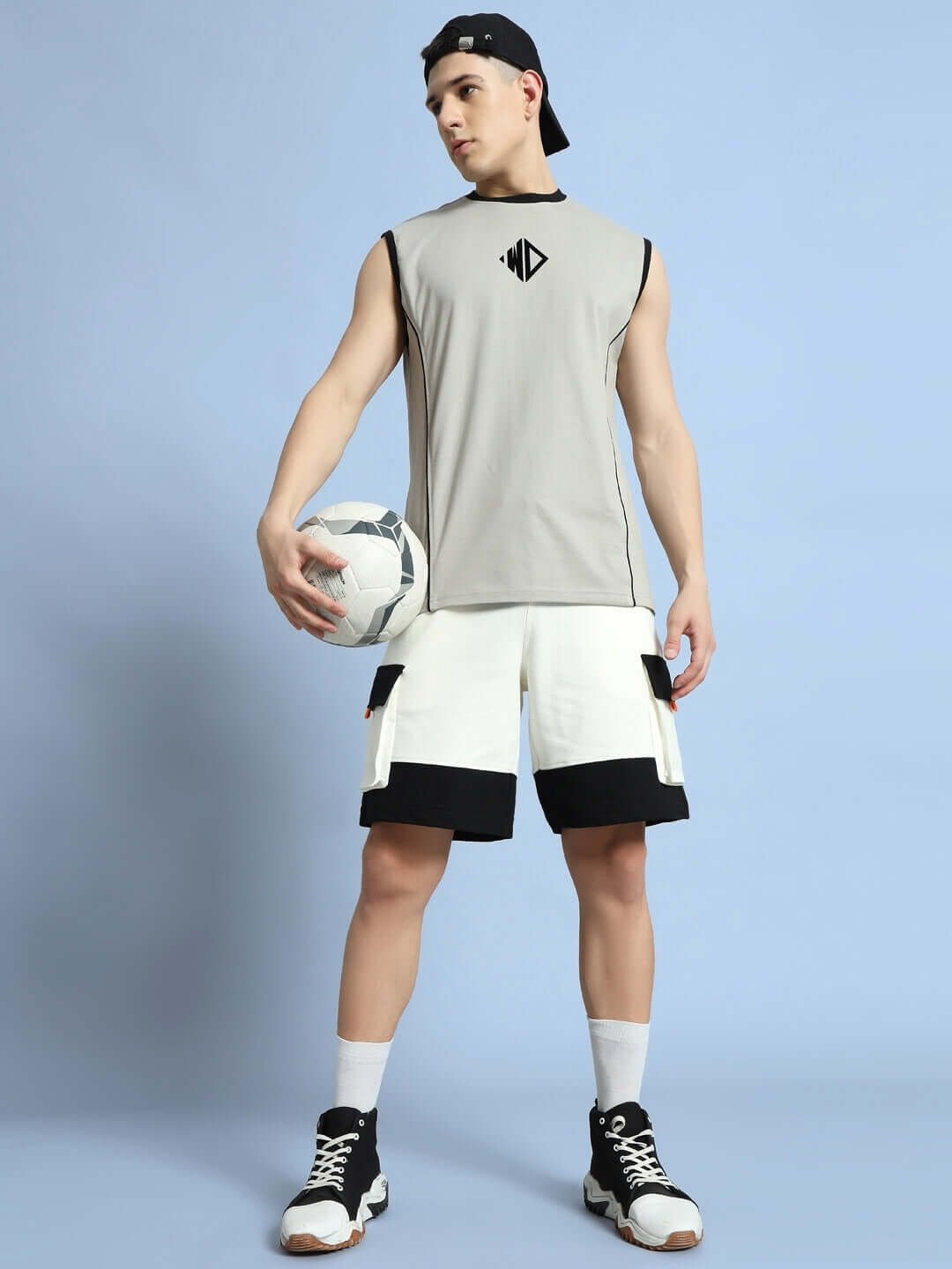 Man wearing off-white and black utility cargo shorts, holding a soccer ball, showcasing casual sporty style against a light blue background.
