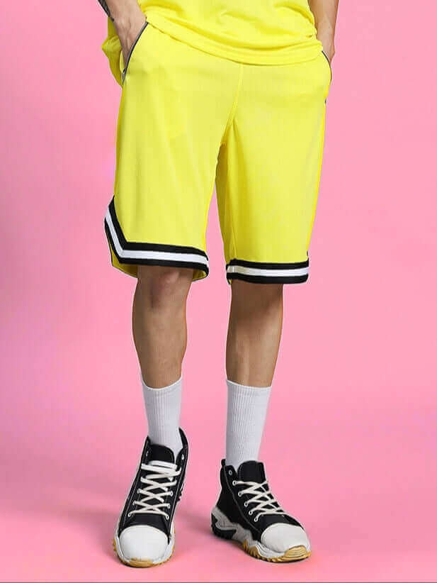 Vigor regular fit yellow shorts with black and white trim, styled with white socks and black sneakers, against a pink background.