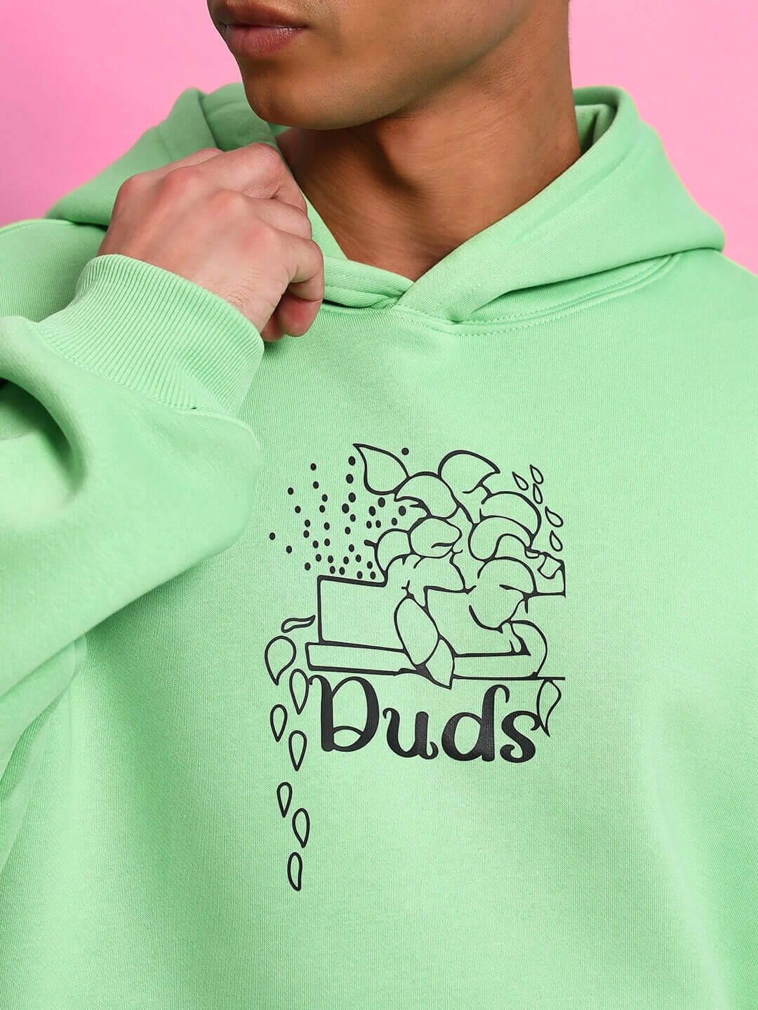 Person wearing VOODO OVERSIZED FLEECE HOODIE GREEN with graphic design, showing detail and texture against pink background.