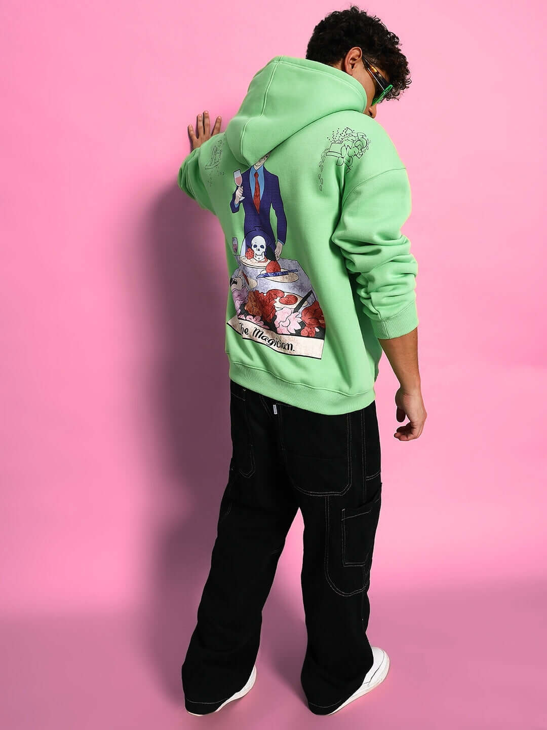 Person wearing VOODO OVERSIZED FLEECE HOODIE GREEN with unique back design, standing against pink background for streetwear style.