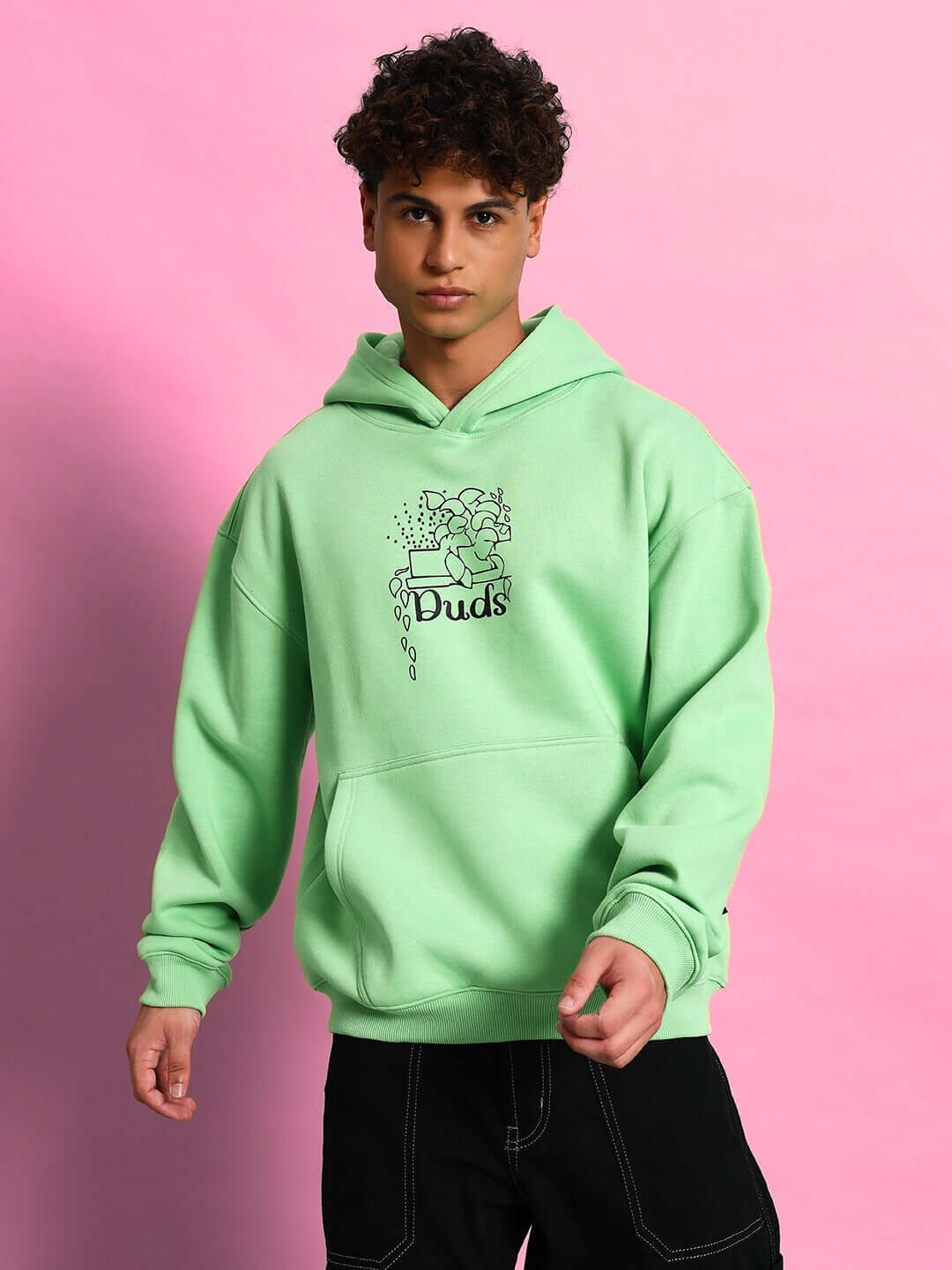 Alt Tag: Man wearing VOODO OVERSIZED FLEECE HOODIE GREEN on pink background, showcasing streetwear style and comfort.