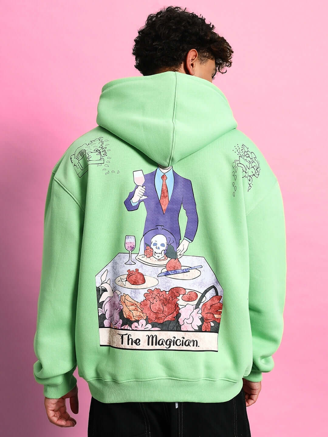 Man wearing VOODO OVERSIZED FLEECE HOODIE GREEN with artistic back print, showcasing unique streetwear design on pink background.