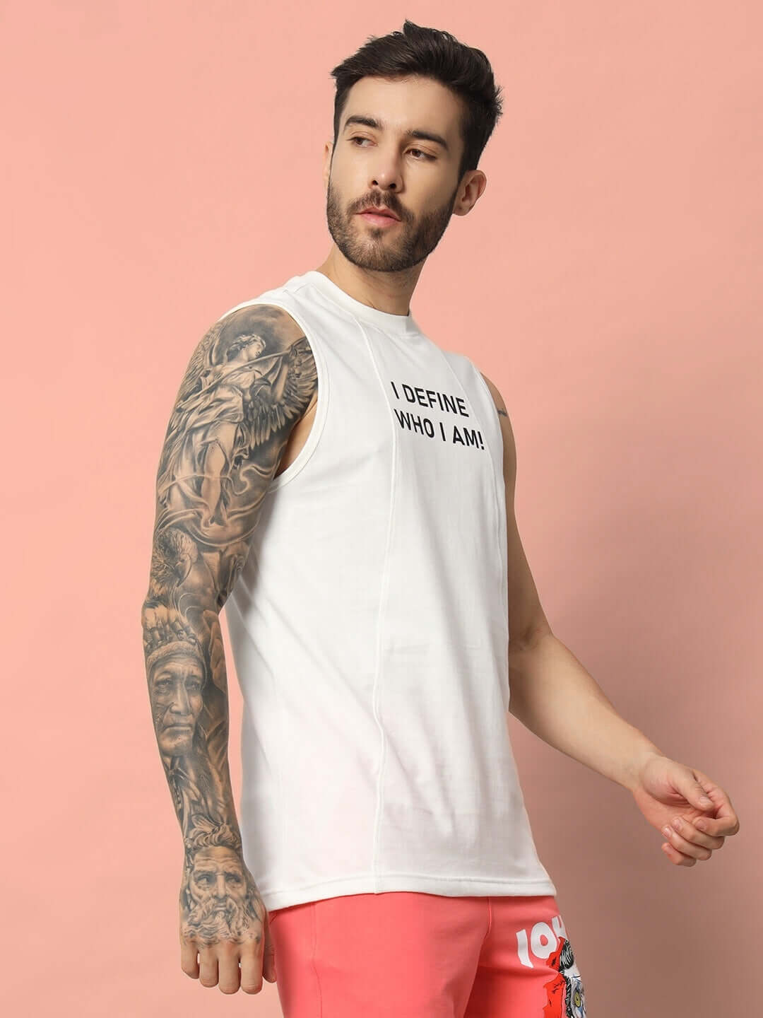 Man wearing WHO I AM GYM T-SHIRT OFF WHITE with tattoos, displaying self-confidence and style in off-white, sleeveless gym apparel.