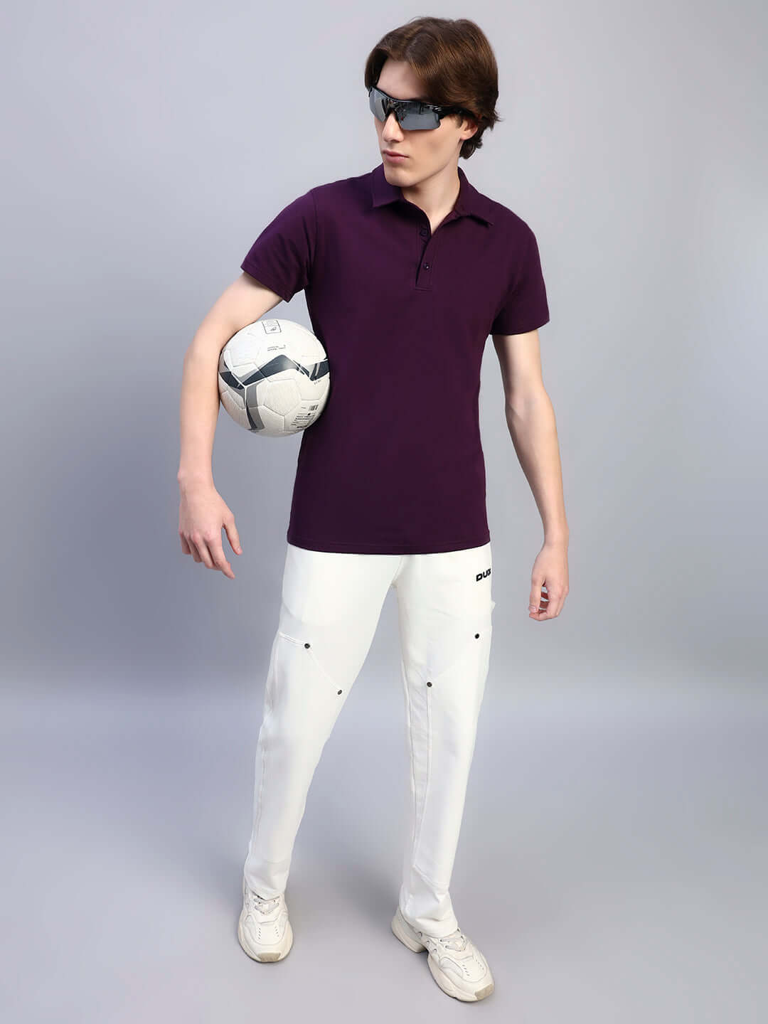 Young man in a WIDEL REGULAR FIT T-SHIRT PURPLE holding a soccer ball, styled with white pants and sunglasses on a neutral background.