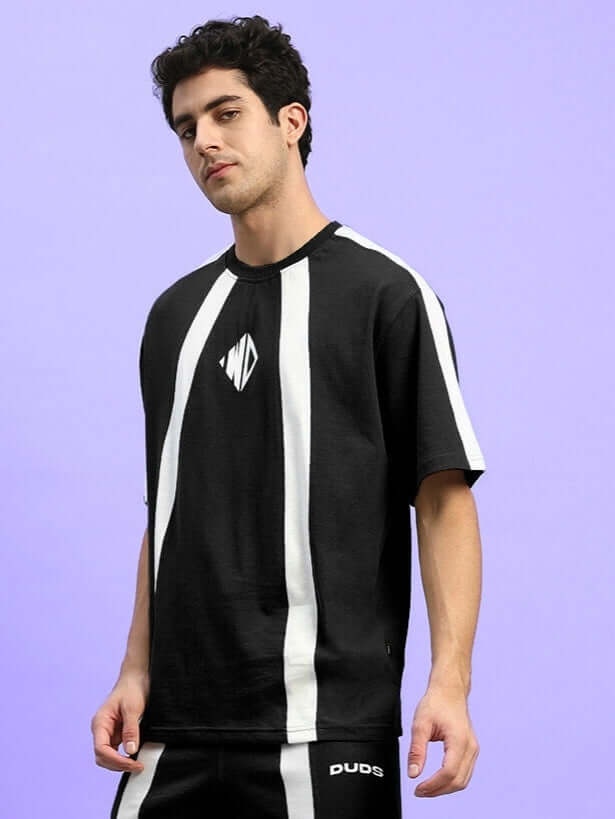 Man wearing WIDELINE OVER-SIZED T-SHIRT BLACK with white stripes, streetwear style, against a purple background.