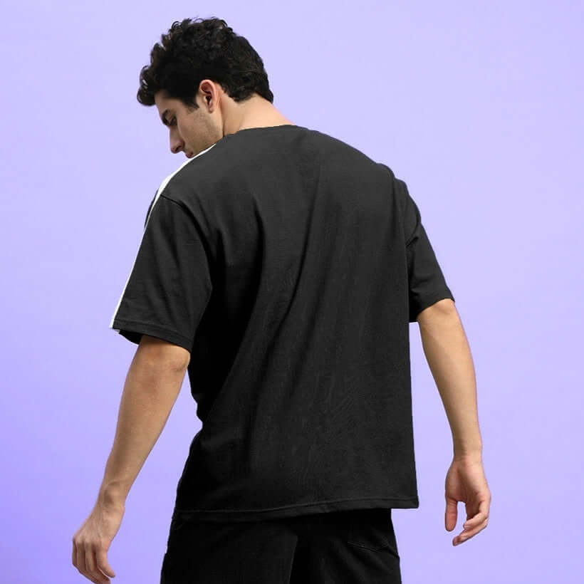Back view of a man wearing a black WIDELINE over-sized t-shirt made from premium cotton French terry, posing against a purple background.