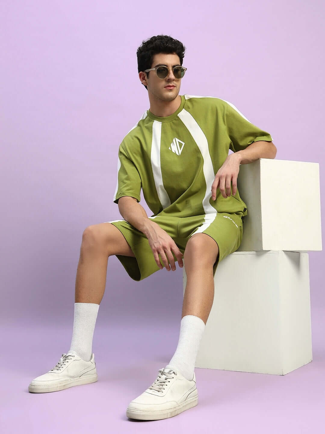 Model wearing WIDELINE REGULAR FIT SHORTS OLIVE GREEN, showcasing casual style in olive green outfit, seated against a lavender background.