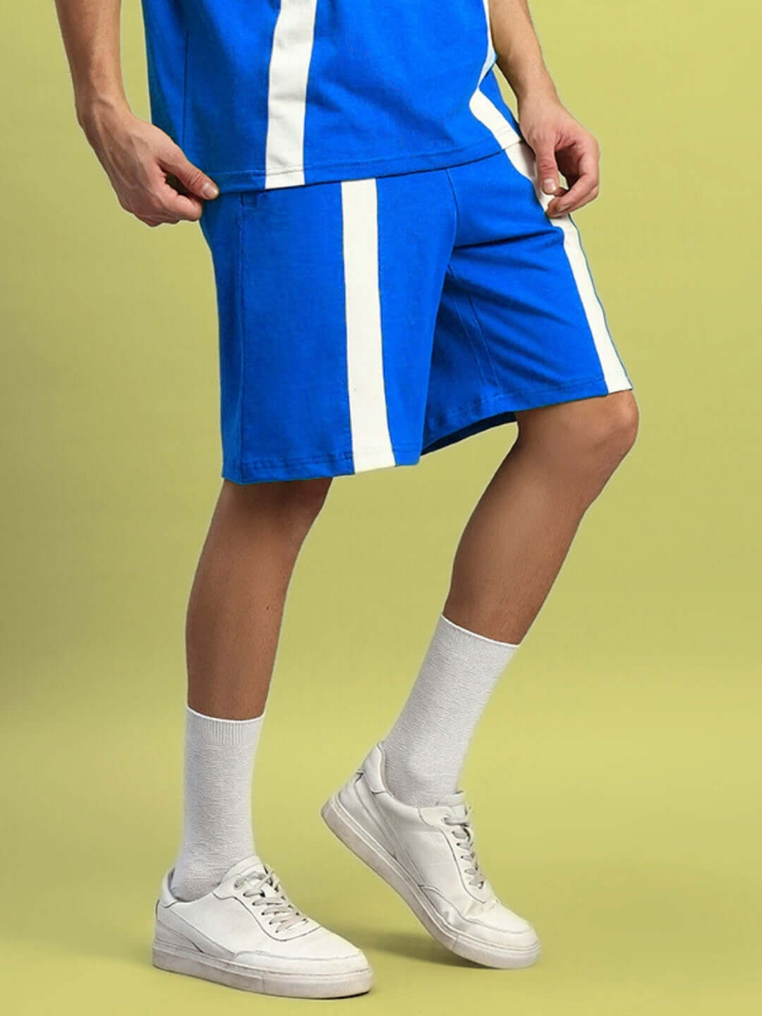 Person wearing WIDELINE REGULAR FIT SHORTS ROYAL BLUE with white stripe, paired with white sneakers, standing on a yellow background.