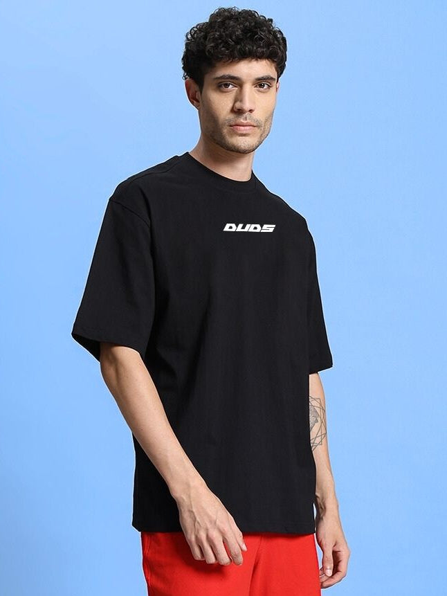 Model wearing WINGED WARRIOR PRINTED OVERSIZED T-SHIRT BLACK with bold graphic, showcasing streetwear style on blue background.