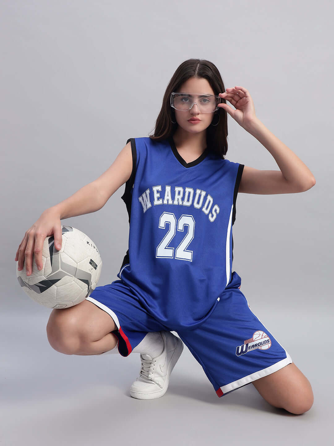 Woman wearing a blue sports jersey and shorts holding a soccer ball, posing confidently on one knee with protective eyewear.