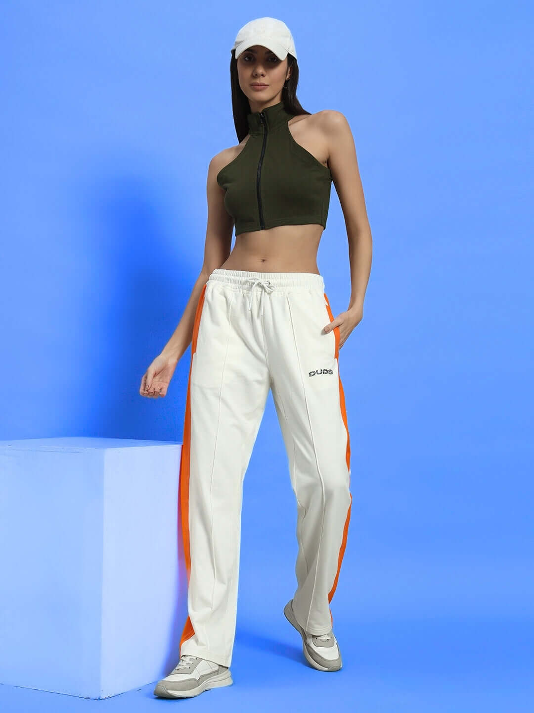 Woman wearing WOMEN'S AURA CROP-TOP - OLIVE GREEN with white pants and cap, standing against a blue background.