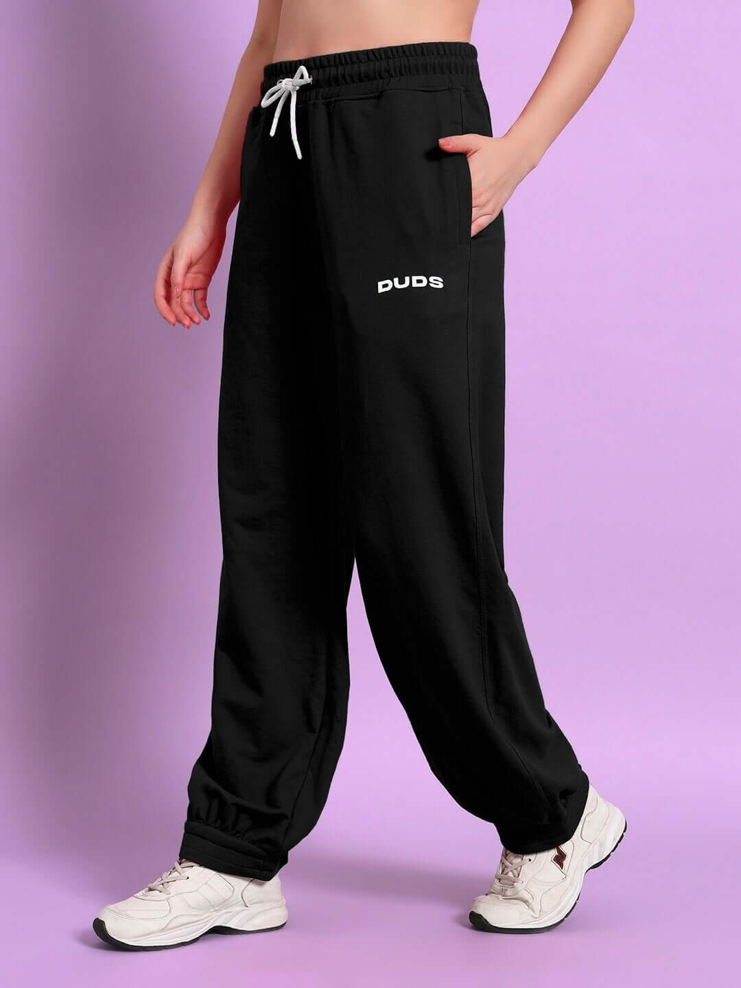 Model wearing women's black relaxed-fit joggers by DUDS with elastic waistband, standing on a pink background.