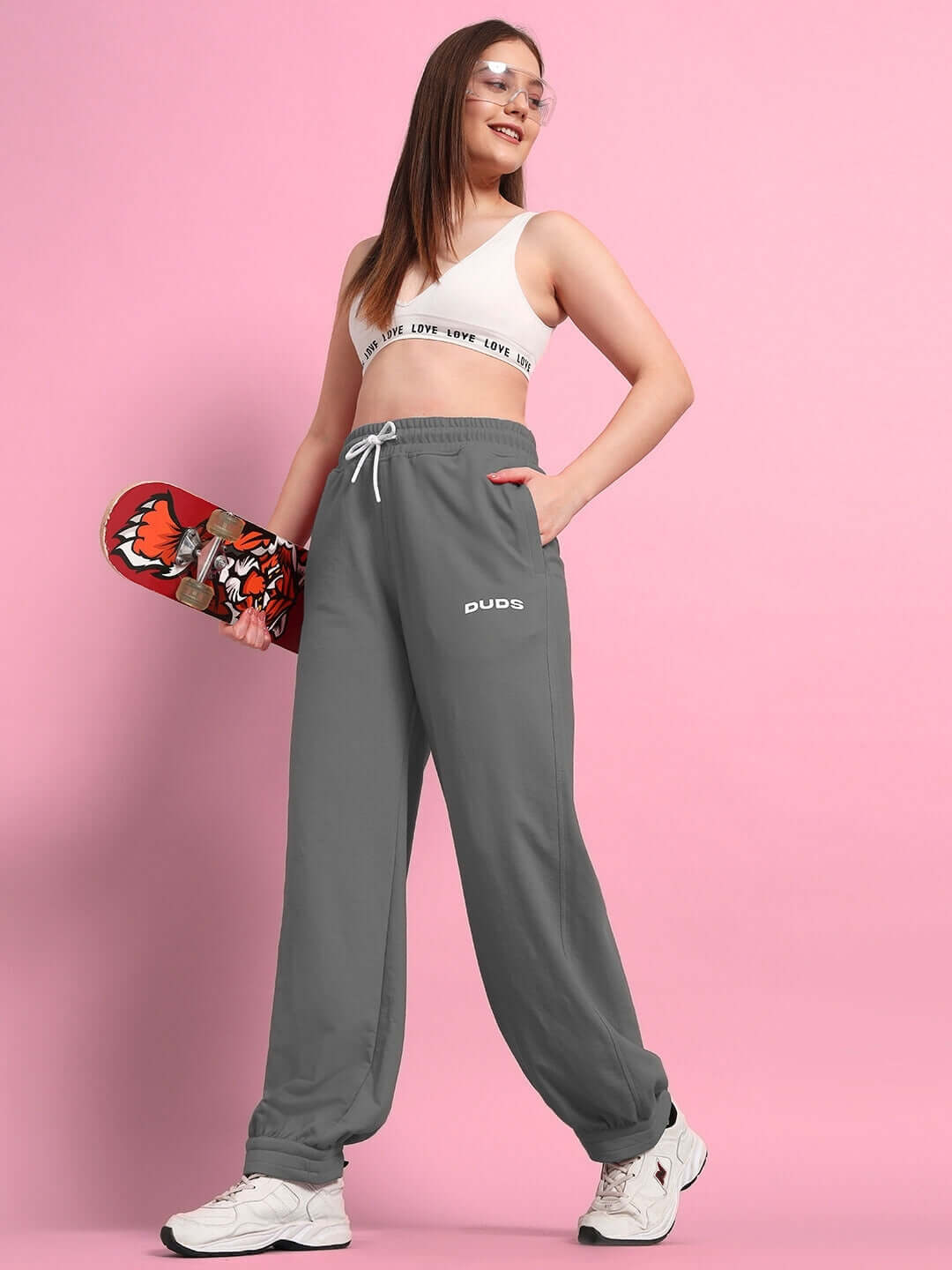 Model wearing WOMENS AUSTRIAN RELAXED-FIT JOGGERS GREY, holding skateboard, stands against pink background.