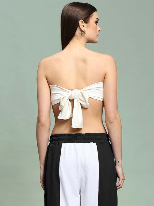 Back view of woman wearing off-white bandeau top with bow tie, paired with black and white pants, on green background.