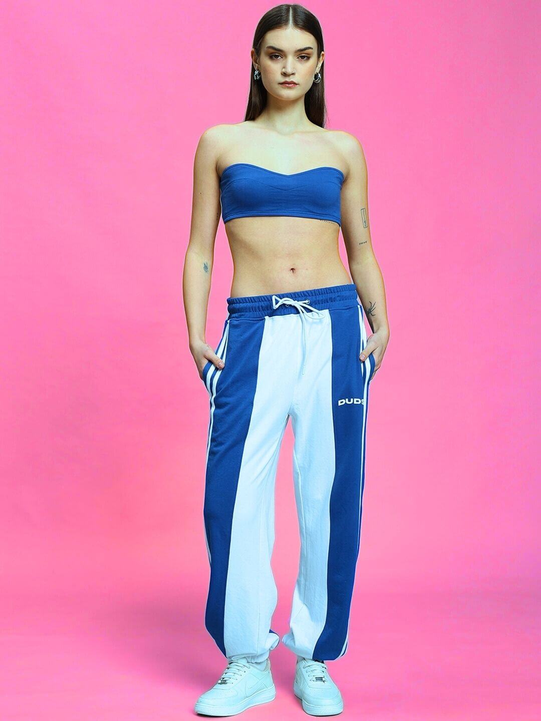 Model wearing Women's Balmy Relaxed Fit Jogger in white and blue, styled with a blue tube top, standing against a pink background.