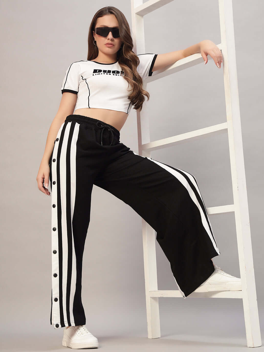 Woman wearing a white Bandit crop-top and black striped pants, styled with sunglasses and white sneakers against a minimalist backdrop.