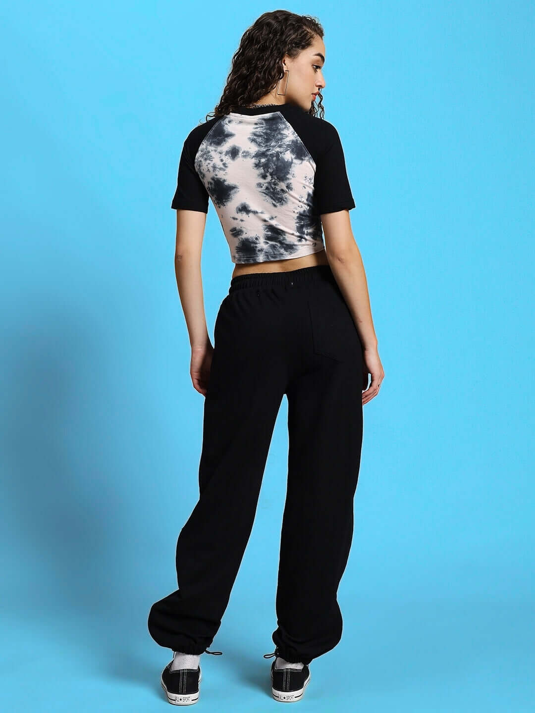 Woman wearing black joggers and black-and-white top, standing with a blue background.