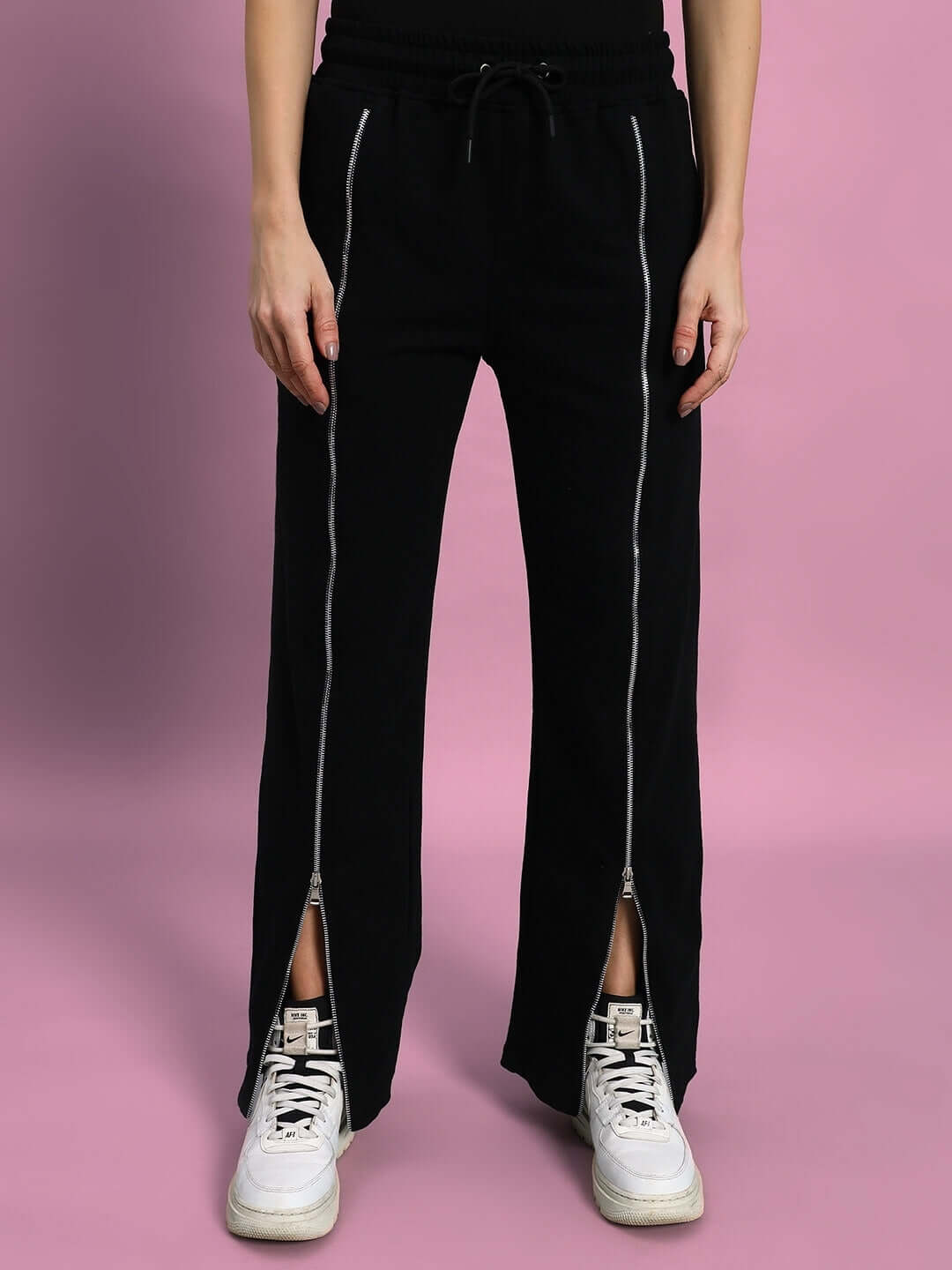 Model wearing black women's joggers with front zipper opening, paired with white sneakers, against a pink background.
