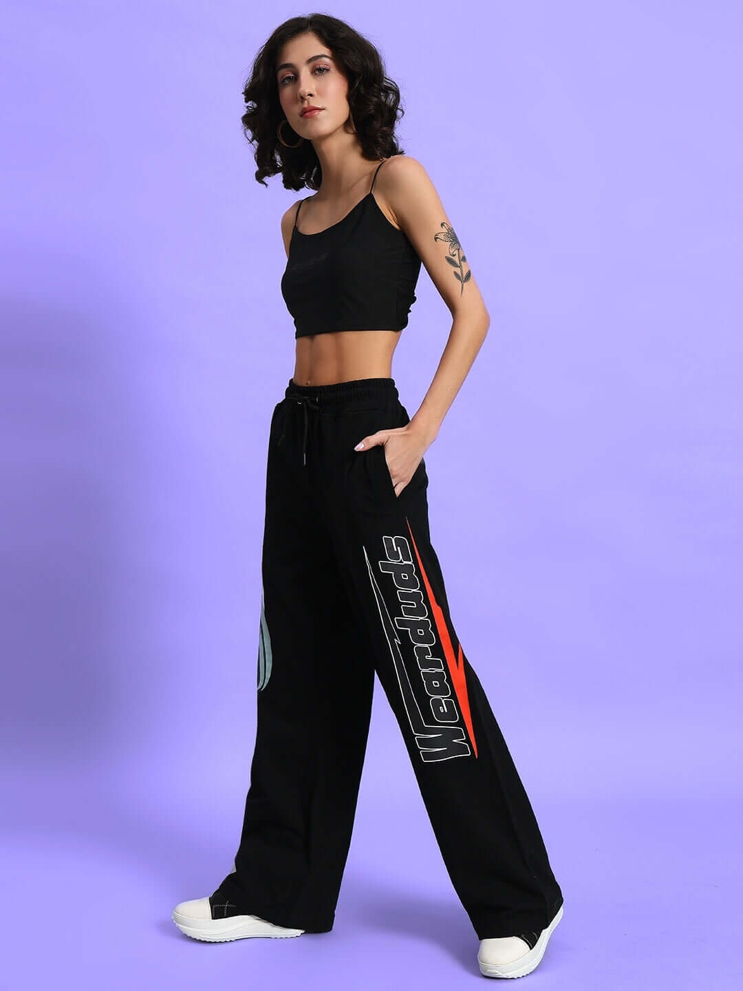Woman wearing black relaxed fit joggers with 'DUDS' logo, standing on a purple background.