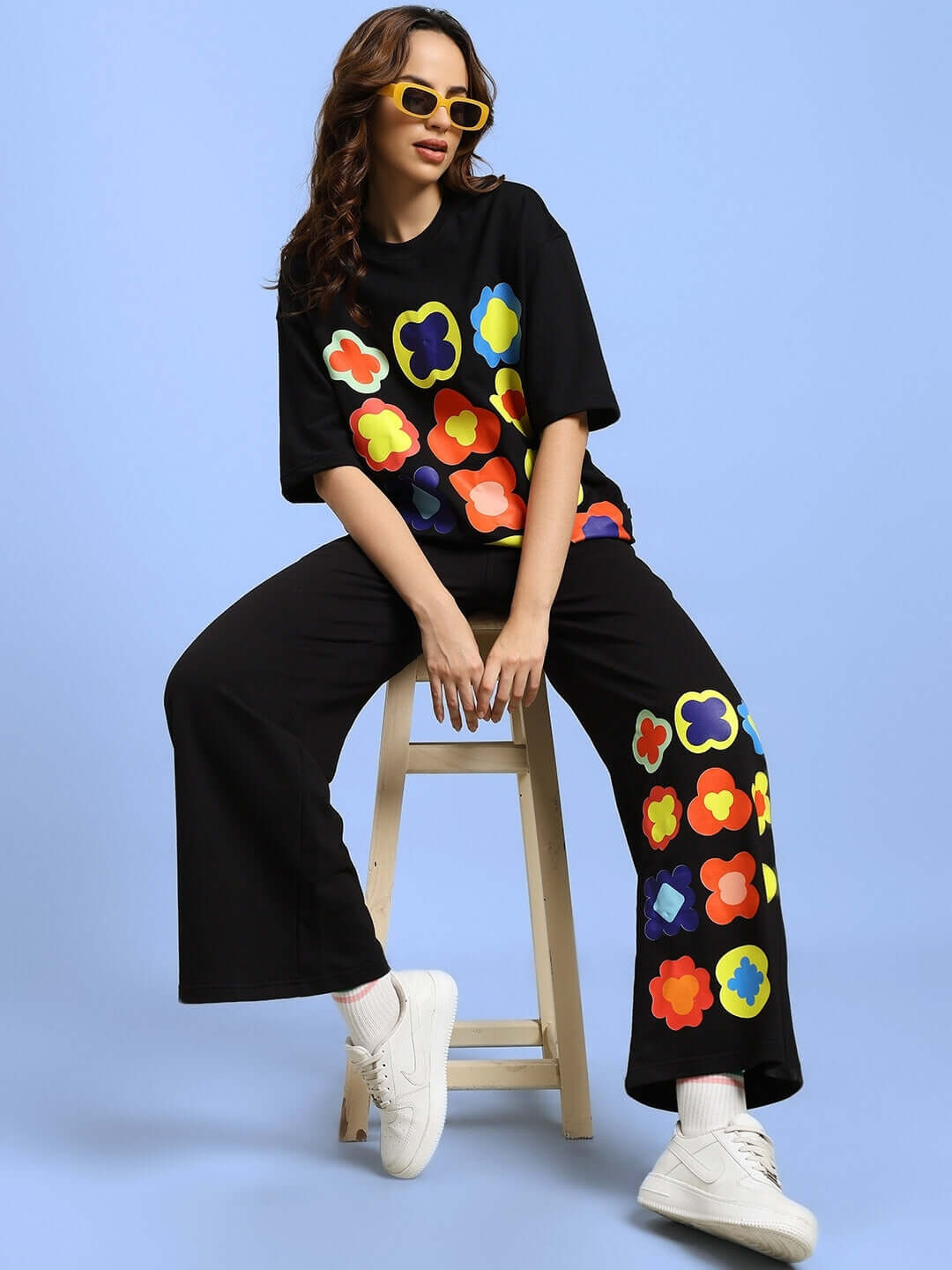 Model wearing WOMEN'S HOLLISTER JOGGERS BLACK with colorful floral motifs seated on a stool against a blue background.