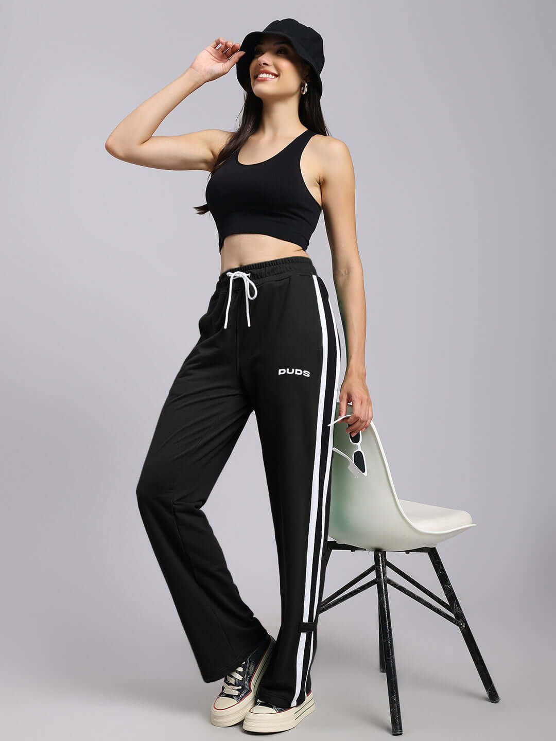 Woman in black relaxed fit joggers and hat, standing and smiling, showcasing DUDS joggers with white stripes.
