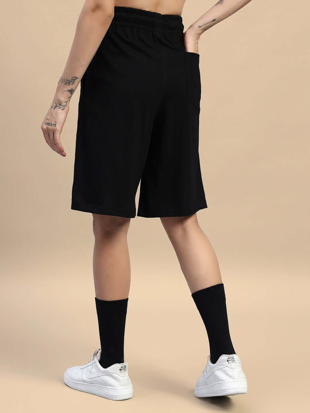 Back view of person wearing black gym shorts with elastic waistband and white sneakers, on beige background.