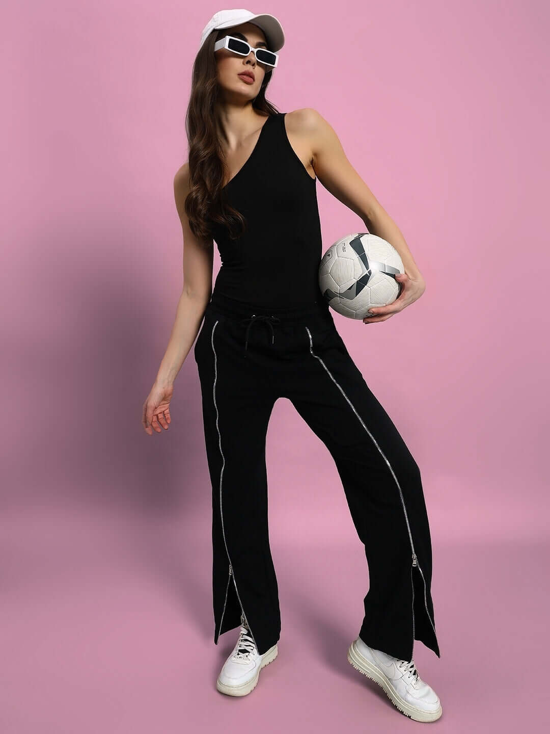 Woman modeling black Brazo front open zipper joggers, holding a soccer ball, wearing a black tank top and white sneakers.