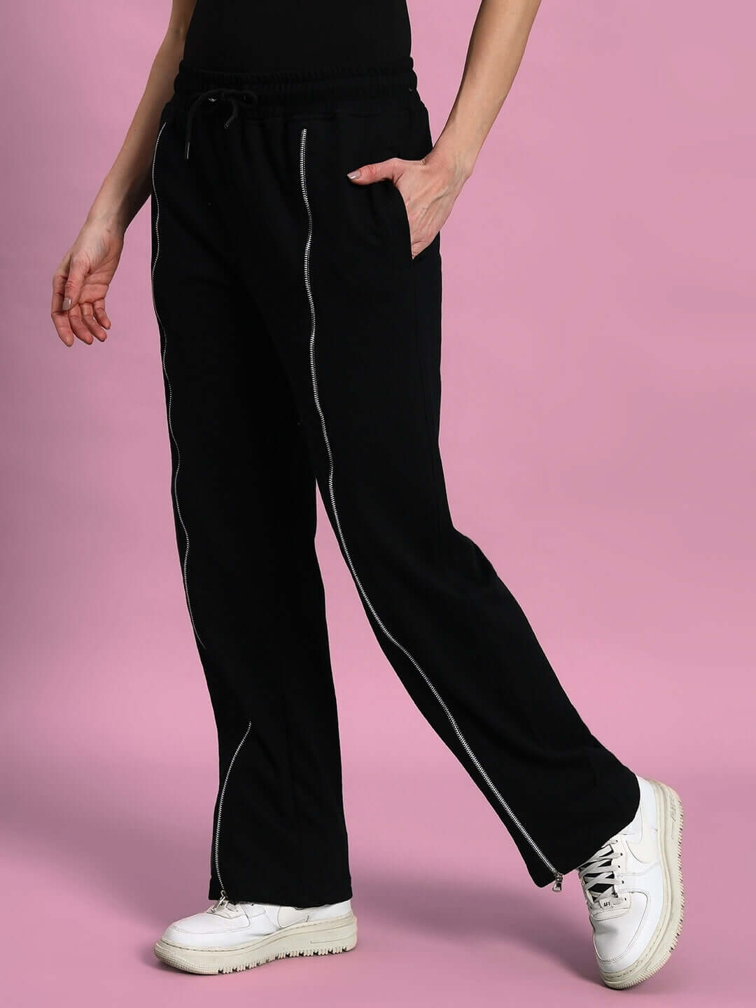 Women's black joggers with front zipper design, worn with white sneakers, against a pink background.