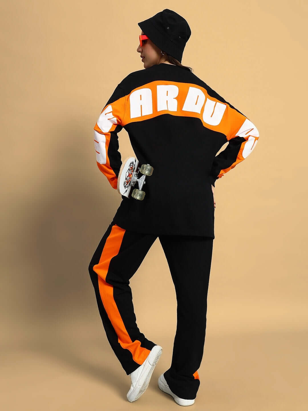 Woman wearing black-orange joggers and matching top, holding a skateboard, against a neutral background.