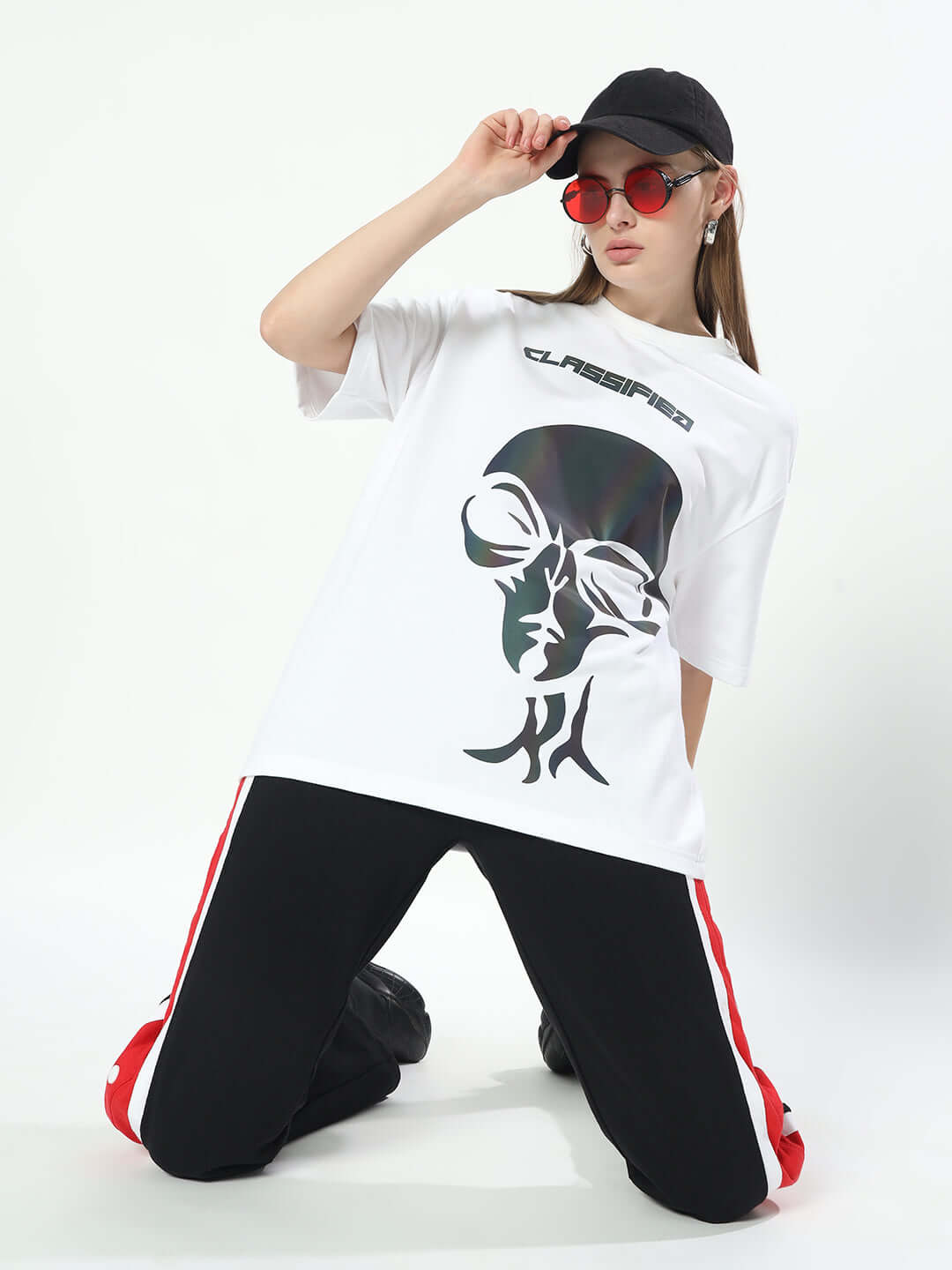 Woman wearing oversized white t-shirt with graphic design, black pants with red stripes, black cap, and red sunglasses, kneeling pose.