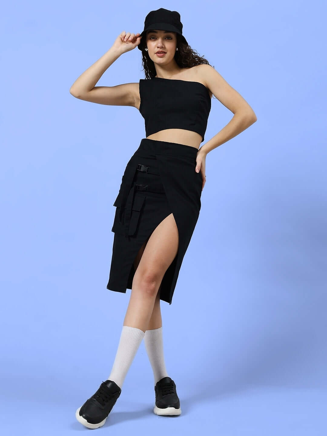 Model wearing women's black Cooksey designer skirt with side slit, paired with a matching black top and cap, showcasing a chic fashion look.