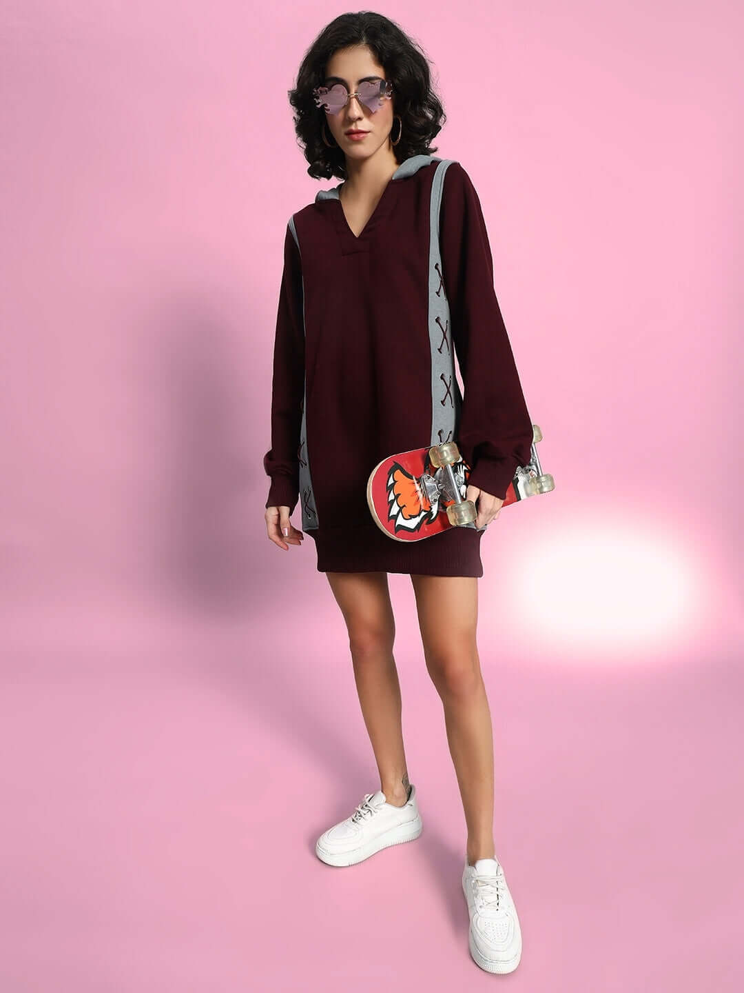 Woman wearing a stylish WOMEN CRISS CROSS FLEECE HOODIE WINE GREY, paired with white sneakers, standing against a pink background.