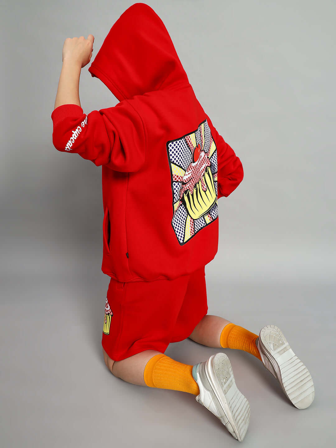 Person wearing WOMEN CUPCAKE FLEECE HOODIE RED showing cupcake graphic on back, kneeling with orange socks and white shoes.