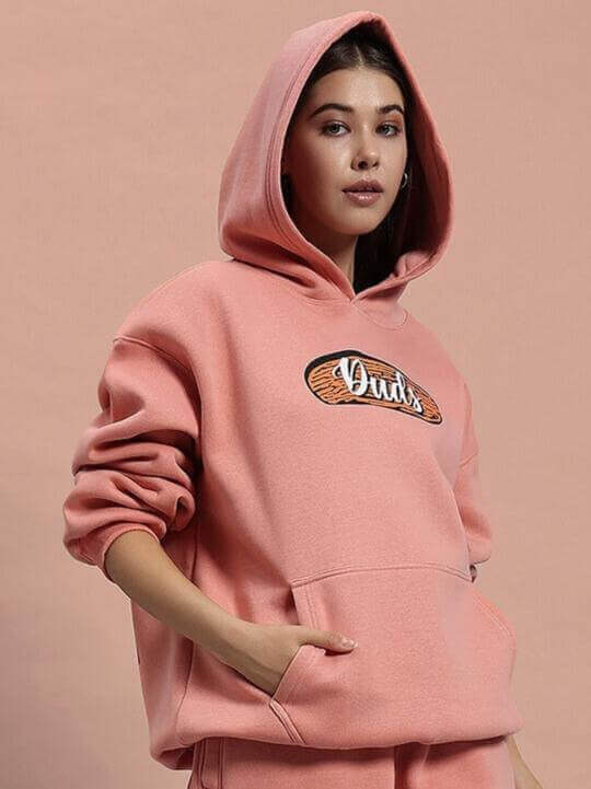 Woman wearing a peach fleece hoodie with a hood, featuring a stylish emblem. Casual and cozy apparel for women.