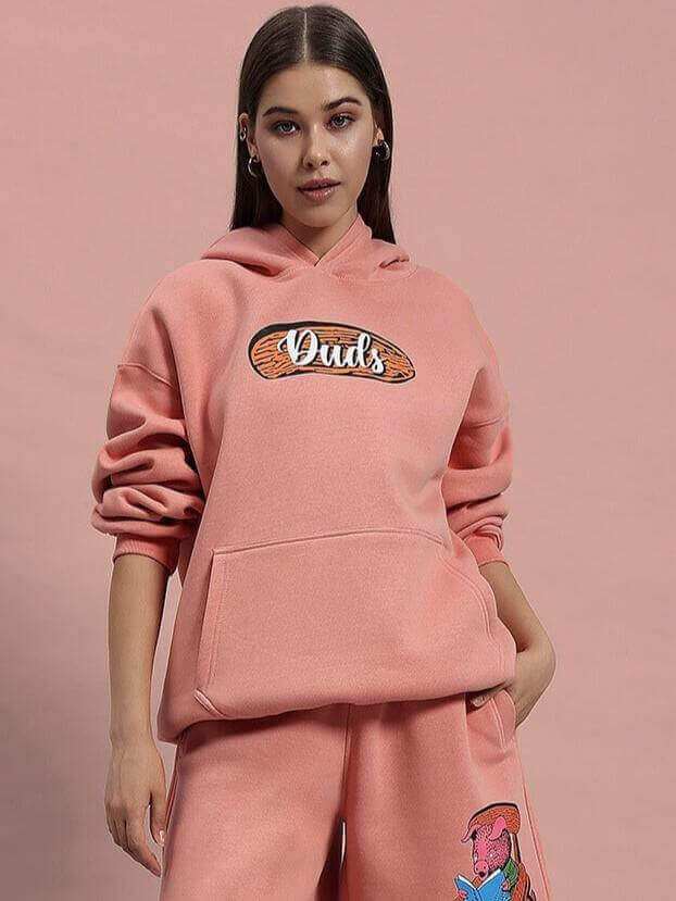 Women's peach fleece hoodie with front pocket and graphic design, styled with matching pants against a peach background.