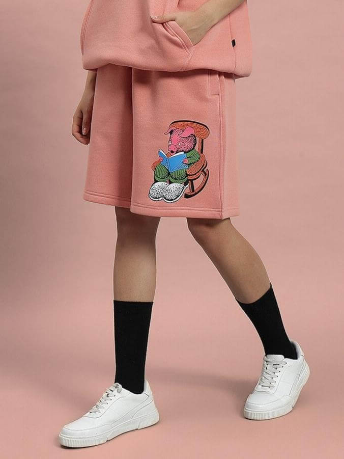 Person wearing WOMEN DELIBERATE FLEECE SHORTS PEACH with bear design, styled with white sneakers and black socks on pink background