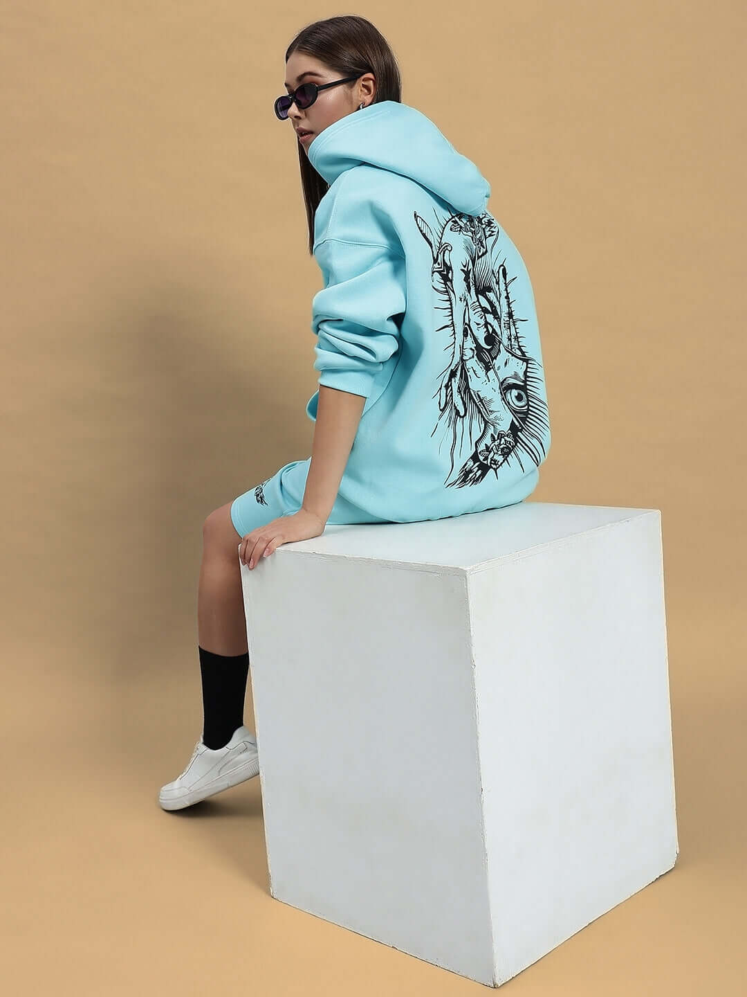 Woman wearing WOMEN DEMON FLEECE HOODIE SKY BLUE sitting on a white cube with stylish design on back.