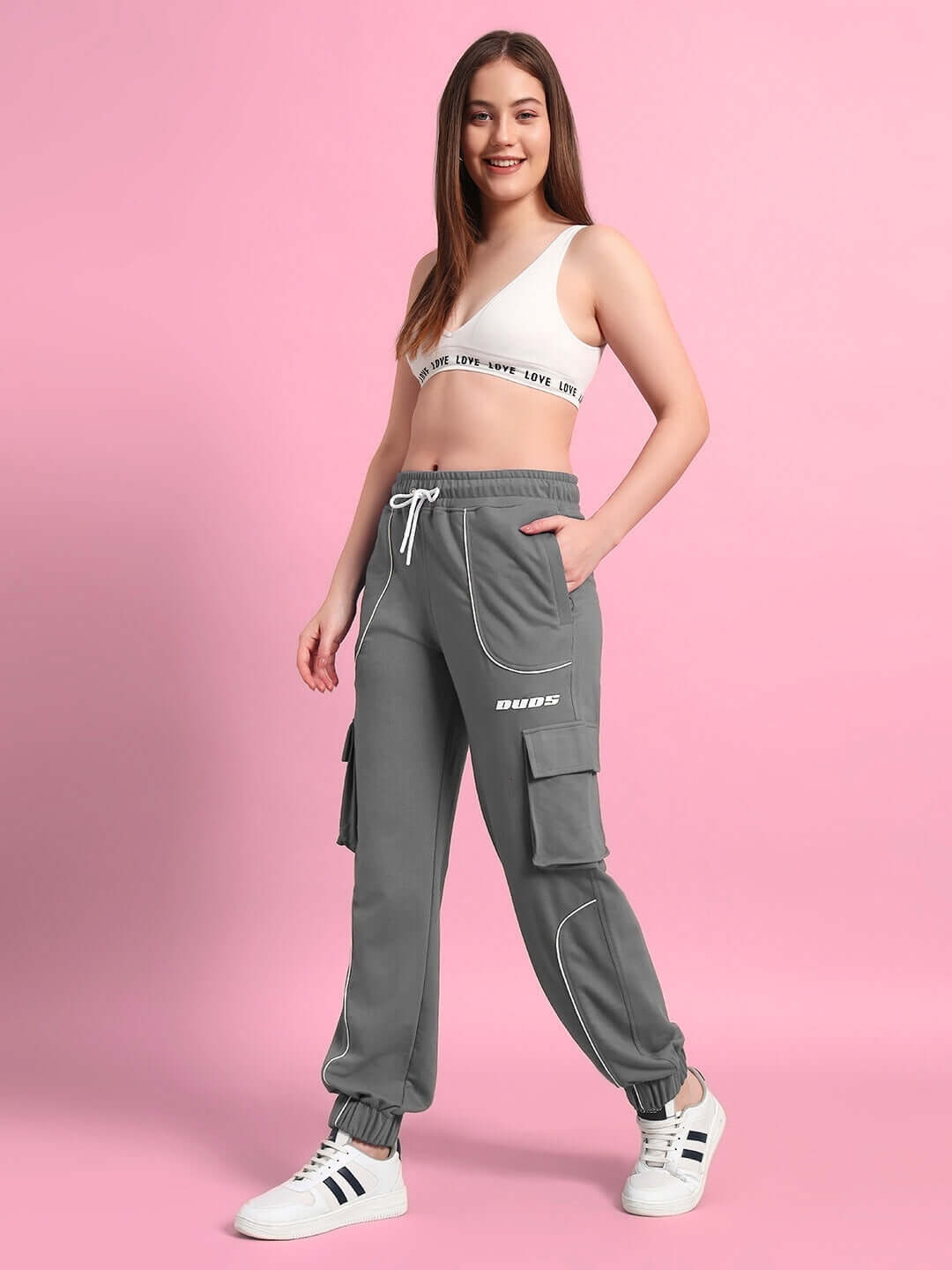 Woman wearing grey relaxed-fit joggers from DUDS standing against a pink background, showcasing casual style and comfort.