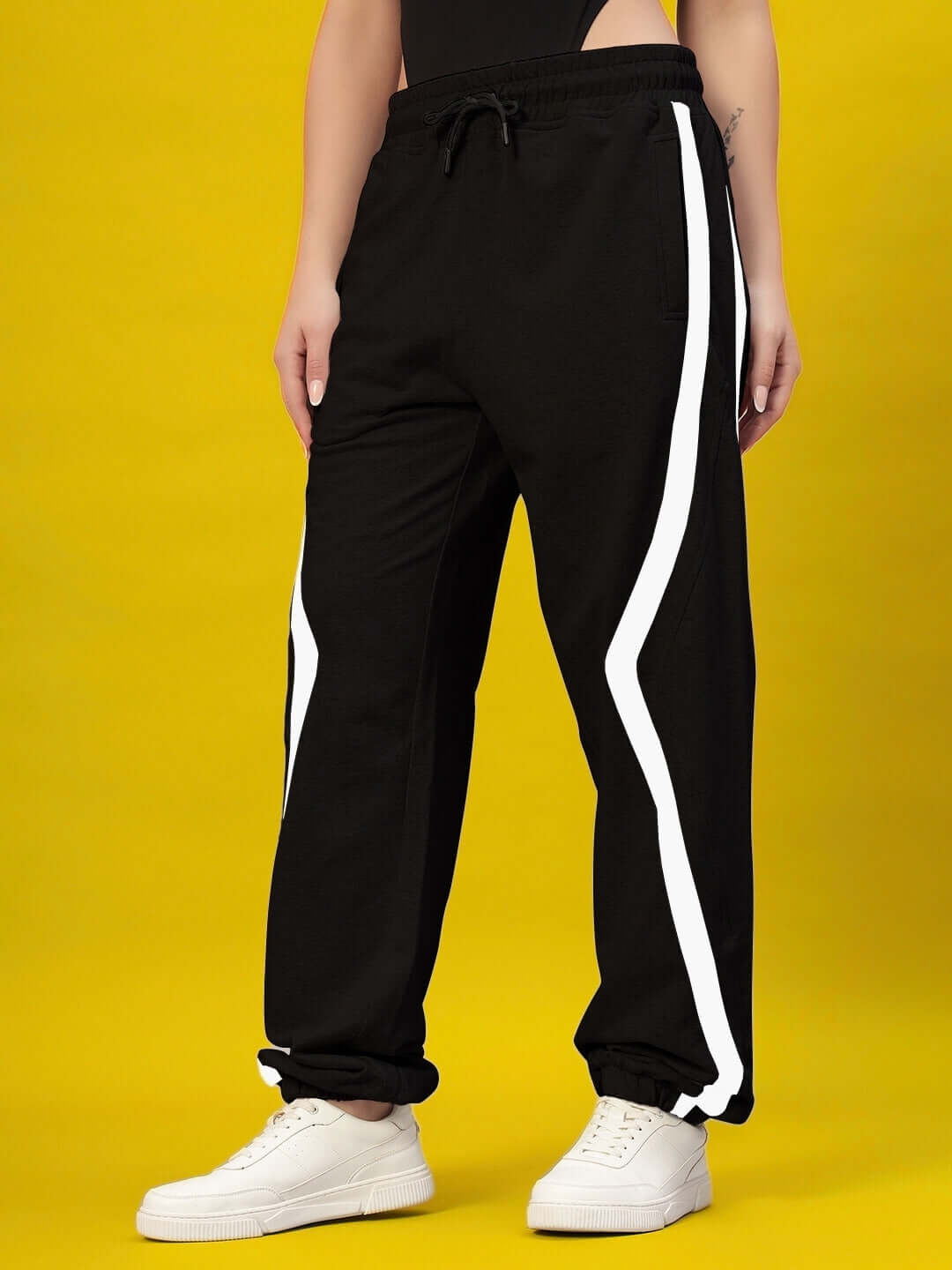 Women's DUDS Frame Joggers in black with white stripes, featuring customizable fit, styled with white sneakers on a yellow background.