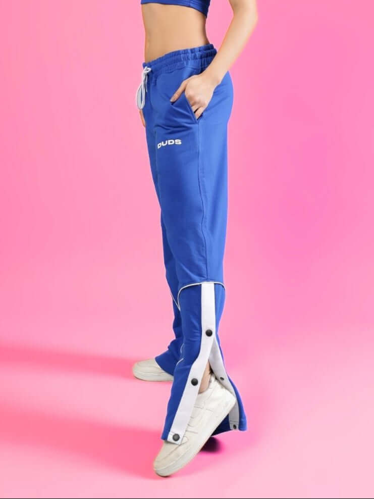 Woman wearing blue Eddie contrast joggers from DUDS, displaying side buttons and pockets against a pink background.