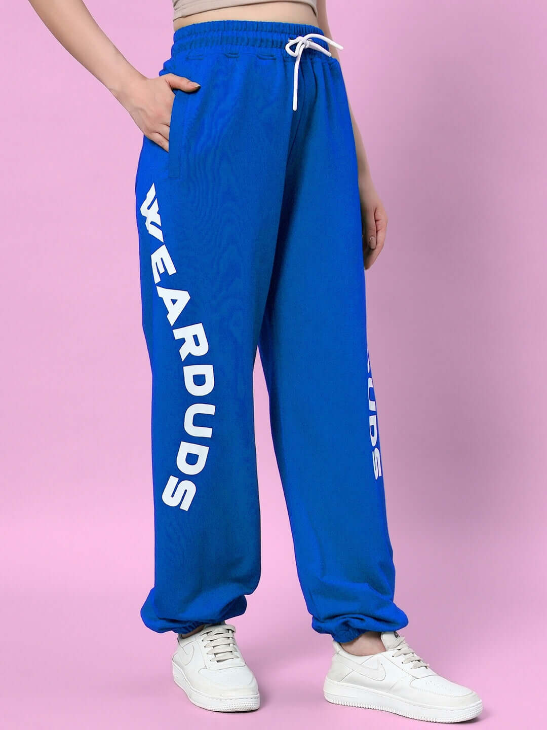 Blue Women's Ellen Relaxed Fit Jogger by DUDS with side pockets, worn with white sneakers against a pink background.
