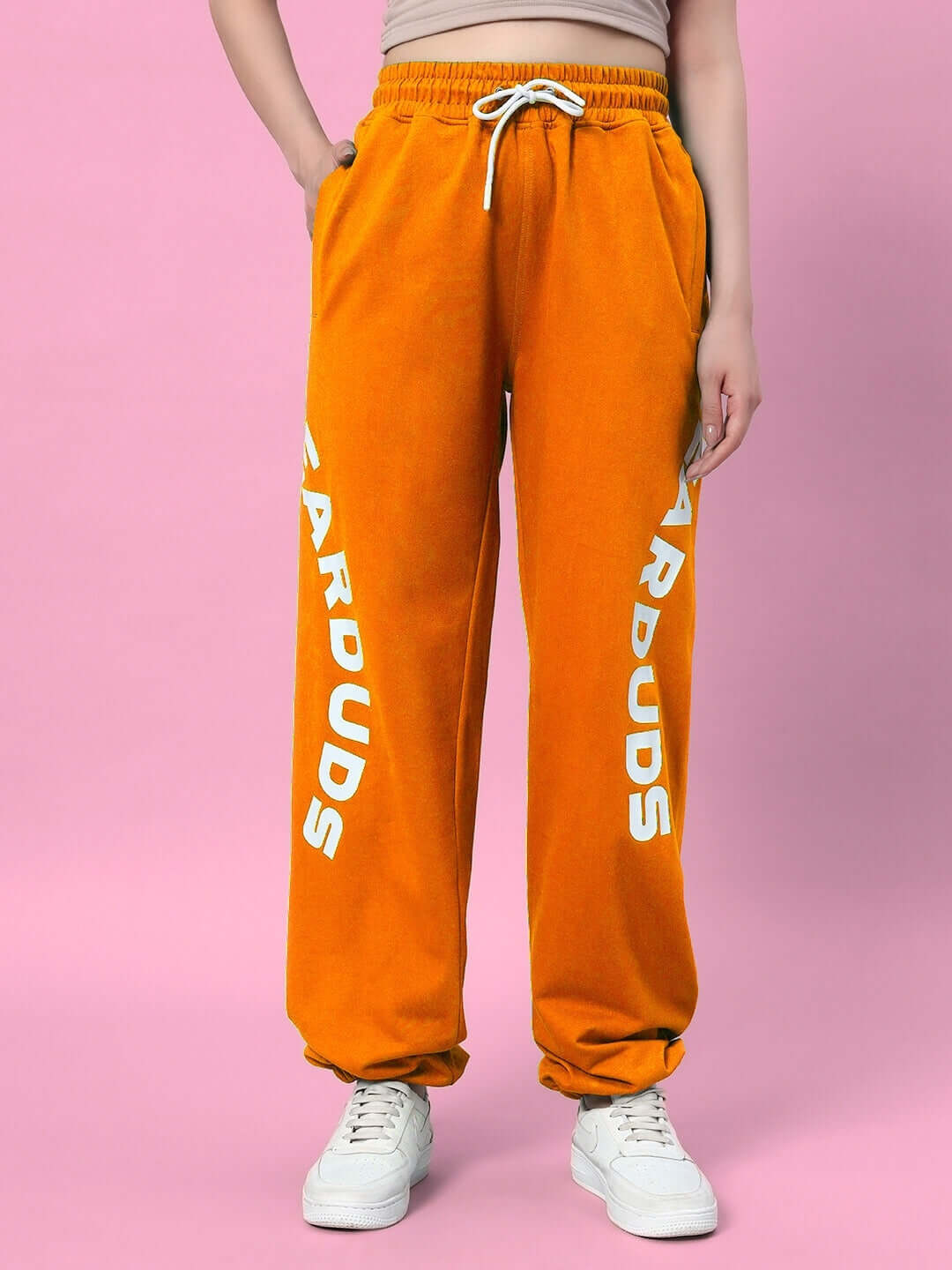 Woman wearing orange joggers with 'DUDS' logo, relaxed fit, tied at the waist, against pink background.