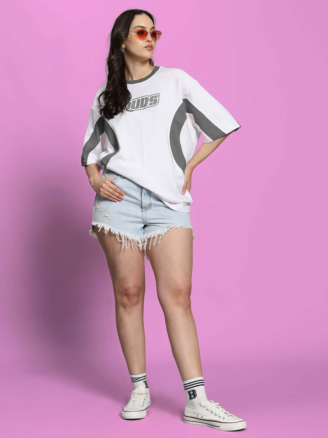 Woman wearing oversized white grey T-shirt with shorts and sneakers, standing against a pink background.