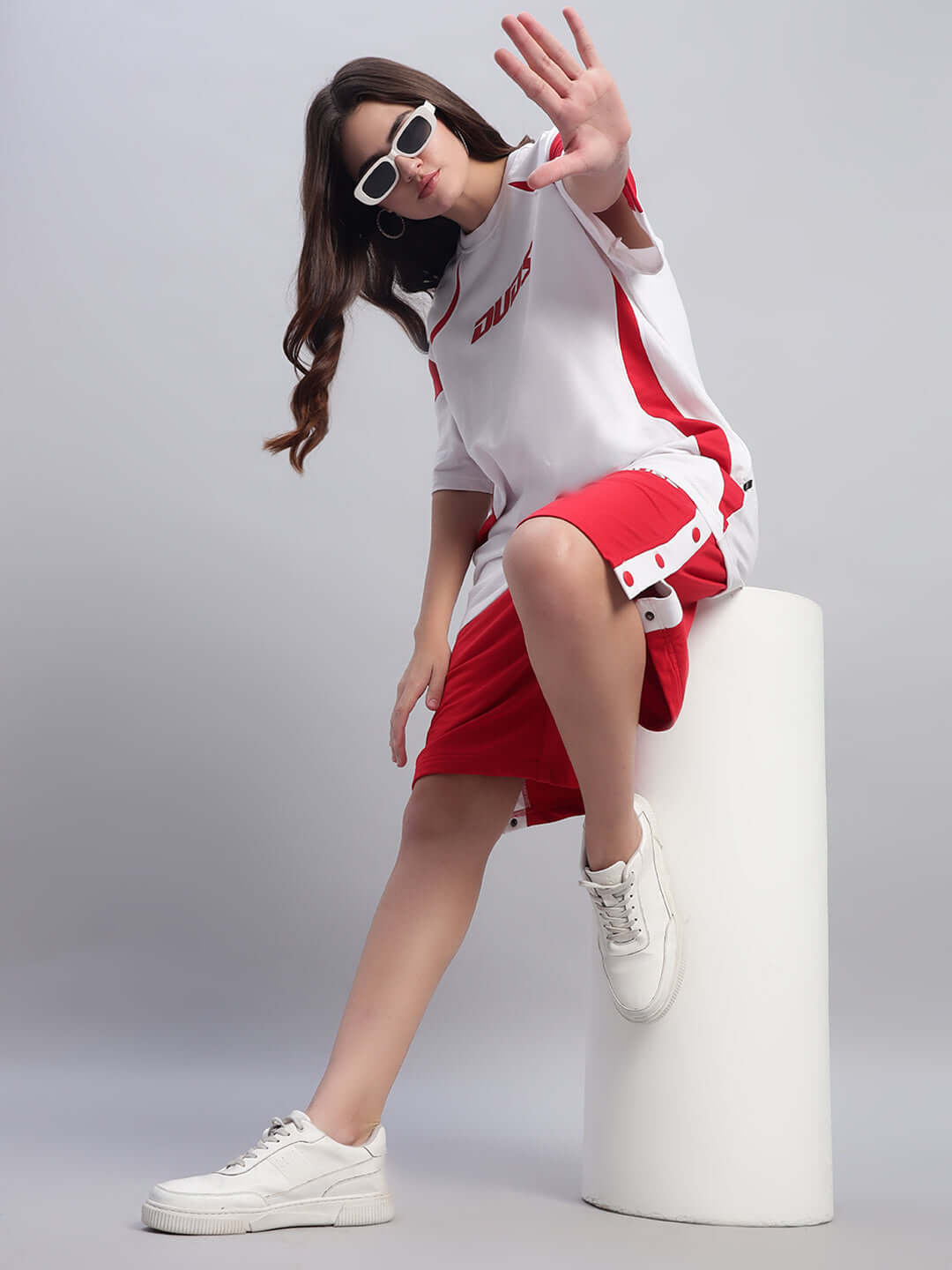 Woman wearing red DUDS shorts and white sneakers, posing casually on a white cylinder, showcasing contemporary athletic style.