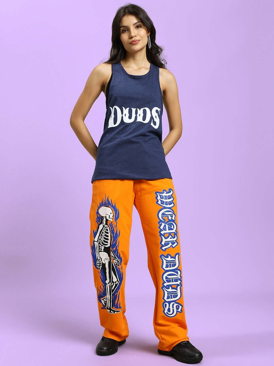Model wearing women's orange joggers with flaming skeleton print, paired with a navy tank top by DUDS, on a purple background.