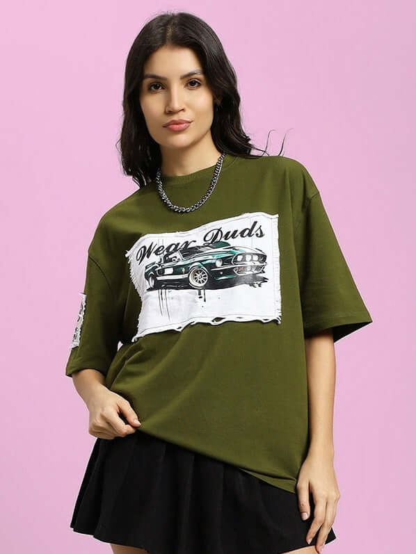 Model wearing olive green oversized T-shirt with Ford Mustang design, made of premium cotton fabric, styled with black skirt on pink background.