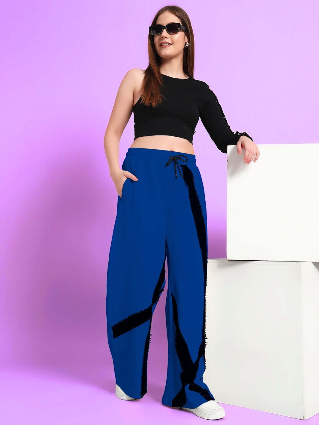 Woman wearing blue frayed textured stripes jogger pants, styled with a black top and sunglasses, standing against a purple backdrop.